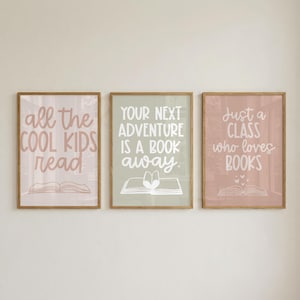 Op de afbeelding: Drie rechthoekige wandposters in houten lijsten. De linker poster is lichtroze met witte tekst all the COOL KIDS read en een boekicoon. De middelste poster is saliegroen met witte tekst YOUR NEXT ADVENTURE IS A BOOK away. en een boekicoon. De rechter poster is oudroze met witte tekst Just a CLASS who loves BOOKS en een boekicoon.