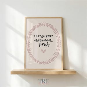 May include: A framed wall art print featuring a light beige background with a dusty pink decorative oval border. The text reads "charge your chromebook, bruh" in black typewriter-style font, accompanied by a small pink heart icon below the text.