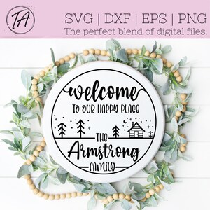 Op de afbeelding: Een rond wit houten bord met zwarte tekst die "Welkom op onze gelukkige plek De familie Armstrong" luidt, met een hut, bomen en sterren rond de tekst.