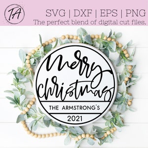 Puede incluir: Un letrero redondo de madera blanco con texto negro que dice "Merry Christmas" con una pancarta negra debajo que dice "The Armstrong's 2021". El letrero est&aacute; rodeado por una guirnalda de hojas verdes y cuentas beige.