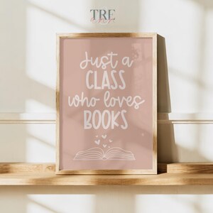 May include: A rectangular wall art print in a light wooden frame featuring a dusty rose background with white text that reads Just a CLASS who loves BOOKS, accompanied by a simple line drawing of an open book and several small floating hearts.