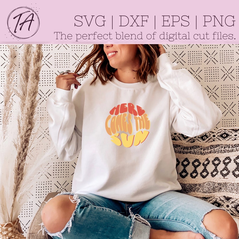 Puede incluir: Sudadera blanca con un gr&aacute;fico retro de un sol con el texto "Here Comes The Sun" en naranja, amarillo y rojo.