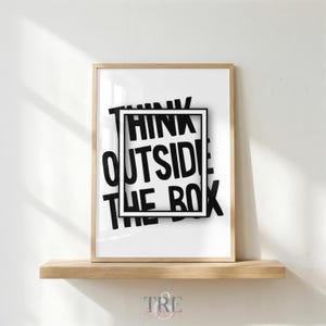 May include: A rectangular wall art print in a light wood frame featuring the black text THINK OUTSIDE THE BOX. The design includes a bold black rectangular border centered over the words, creating a visual representation of the phrase.
