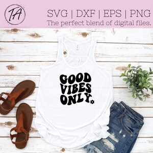 Op de afbeelding: Een wit tanktop met zwarte tekst die "GOOD VIBES ONLY" luidt in een retro lettertype. Het tanktop ligt op een wit houten oppervlak.