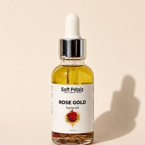 May include: A clear glass dropper bottle containing golden facial oil with visible dried flower petals at the bottom. The white label reads Soft Pétalz, Clean. Organic. Gentle. ROSE GOLD Facial Oil, and 1 Oz USA, with a red rose illustration.