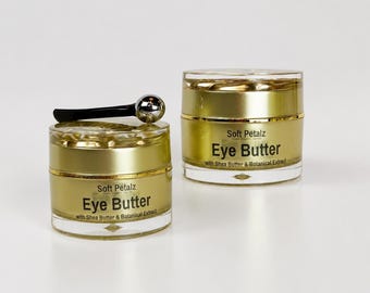 May include: Two cylindrical jars of Soft Petalz Eye Butter with Shea Butter & Botanical Extract. The jars have gold-toned lids and bases with clear midsections. A small black and silver metal applicator tool rests on the lid of the left jar.