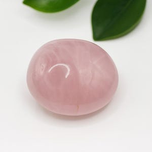 Peut inclure: Une pierre de quartz rose lisse et ovale, de couleur rose pâle, est posée sur une surface blanche. La pierre est le point central, avec un éclat brillant. Des feuilles vertes sont en arrière-plan.
