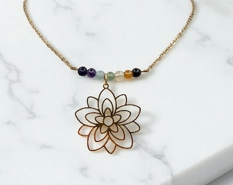 May include: A gold-colored necklace featuring a lotus flower pendant. The necklace has a delicate chain and a row of colorful beads. The pendant is a detailed outline of a lotus flower. The necklace is displayed on a white marble surface.