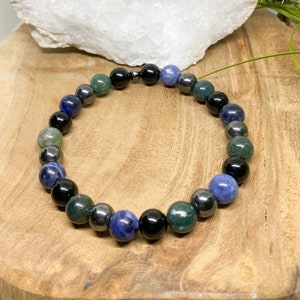 May include: A beaded bracelet featuring a mix of polished gemstones. The bracelet includes black, dark green, blue, and silver-gray beads. The bracelet is displayed on a wooden surface with a white crystal geode in the background.