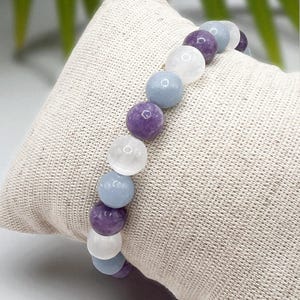 May include: A beaded bracelet featuring a mix of purple, light blue, and white round gemstones. The bracelet is displayed on a beige fabric surface, showcasing the colors and textures of the stones. The bracelet is a piece of jewelry.