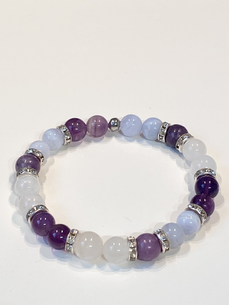 May include: A beaded bracelet featuring a mix of purple, white, and light blue gemstones. The bracelet includes silver-toned spacers with clear crystal accents. The gemstones are arranged in a circular pattern, creating a stylish accessory.