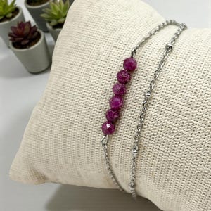 Op de afbeelding: Een zilveren kettingarmband met een rij gefacetteerde, dieproze edelstenen. De armband ligt op een beige, getextureerd kussen. Kleine vetplanten in potten staan op de achtergrond.