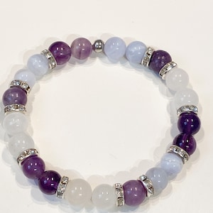May include: A beaded bracelet featuring a mix of purple, white, and light blue gemstones. The bracelet has silver-colored spacers with clear rhinestones. The gemstones are round and vary in size, creating a visually appealing accessory.