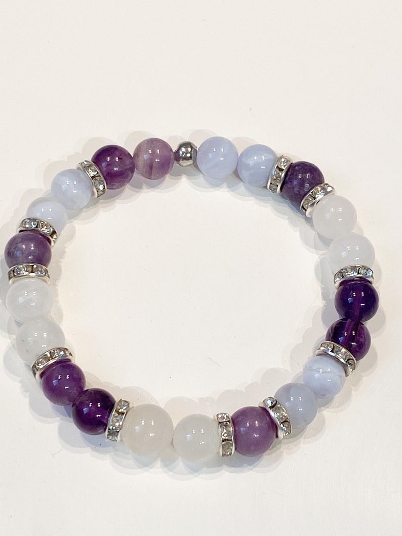 May include: A beaded bracelet featuring a mix of purple, white, and light blue gemstones. The bracelet has silver-colored spacers with clear rhinestones. The gemstones are round and vary in size, creating a visually appealing accessory.