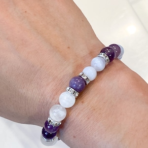 May include: A beaded bracelet featuring alternating purple and white gemstone beads. The bracelet is accented with silver-colored spacers encrusted with small, clear crystals. The bracelet is worn on a wrist.