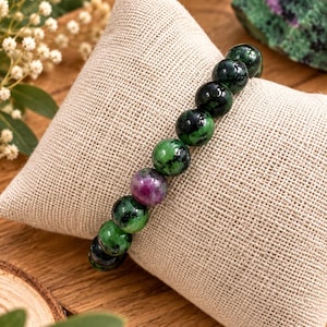 Pode incluir: Uma pulseira de contas com pedras redondas e polidas em tons de verde floresta profundo e preto, com uma conta esférica proeminente de cor magenta-rosa vibrante posicionada perto do centro da banda elástica.