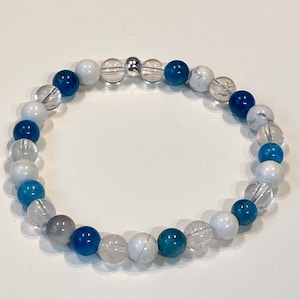 May include: A blue and white beaded bracelet with a silver charm. The bracelet is made with clear quartz, blue apatite, and white howlite beads.