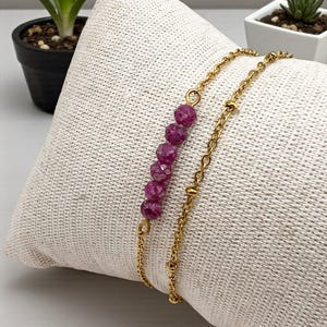 Può includere: Un braccialetto a catena color oro con una fila di sei pietre preziose rosa intenso sfaccettate. Il braccialetto è esposto su una superficie bianca e strutturata. Il braccialetto ha due catene.