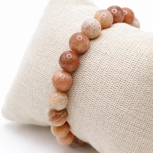 May include: A beaded bracelet featuring round, polished beads in shades of peach, tan, and brown. The bracelet is displayed on a beige, textured fabric. The beads have a natural, marbled appearance, suggesting they are made of stone or a similar material.