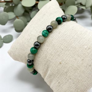 May include: A bracelet with a mix of green, grey, and black beads. The bracelet is displayed on a beige, textured cushion. The beads are round and vary in size, creating a visually appealing accessory.