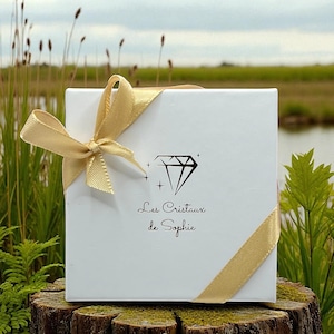 May include: A white gift box tied with a gold satin ribbon and bow. The box features a diamond graphic and the text "Les Cristaux de Sophie" in black script. The box is sitting on a tree stump.