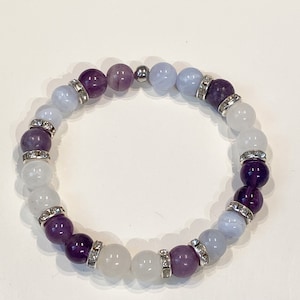 May include: A beaded bracelet featuring a mix of purple, white, and light blue gemstones. The bracelet has silver-colored spacers with clear rhinestones. The beads are strung on an elastic band, creating a stretchable design. The bracelet is a piece of jewelry.