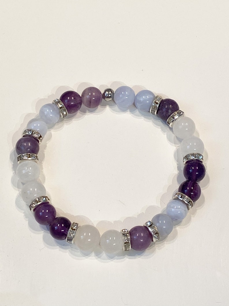 May include: A beaded bracelet featuring a mix of purple, white, and light blue gemstones. The bracelet has silver-colored spacers with clear rhinestones. The beads are strung on an elastic band, creating a stretchable design. The bracelet is a piece of jewelry.