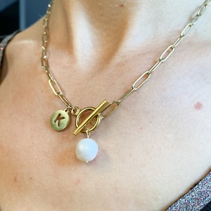 Può includere: Una collana a catena dorata con chiusura a levetta e un ciondolo di perla. Il ciondolo è attaccato a un piccolo disco dorato con la lettera "K" sopra.