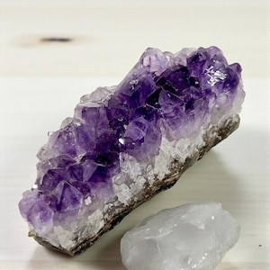 Peut inclure: Un amas de cristaux d'améthyste violet foncé avec une pierre de quartz blanche. L'améthyste est d'un riche violet foncé, et le quartz est blanc laiteux. Les cristaux sont sur une surface de couleur claire.