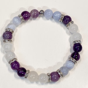 May include: A beaded bracelet featuring a mix of purple, lavender, light blue, and white gemstones. The bracelet includes silver-toned spacers with clear rhinestones. The beads are strung on an elastic band, creating a stretch bracelet.