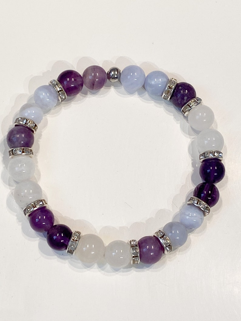 May include: A beaded bracelet featuring a mix of purple, lavender, light blue, and white gemstones. The bracelet includes silver-toned spacers with clear rhinestones. The beads are strung on an elastic band, creating a stretch bracelet.