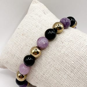 Può includere: Un braccialetto di perline con un mix di perline dorate, nere e viola. Il braccialetto è esposto su una superficie strutturata di colore neutro. Le perline sono rotonde e di dimensioni variabili, creando un accessorio visivamente accattivante.