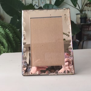 May include: A rectangular, vintage-style photo frame with a scalloped edge. The frame is made of mirrored glass, reflecting the surrounding environment. The back of the frame is a light brown color, and the frame is standing upright on a white surface.