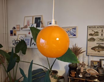 May include: A bright orange glass pendant light fixture hangs from a white ceiling fixture. The light is round and has a smooth, glossy finish. The light is surrounded by green plants.