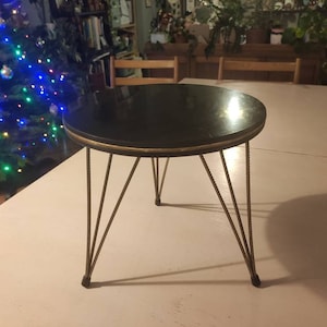 Può includere: Un tavolino rotondo nero con un bordo dorato attorno al bordo del piano. Il tavolo ha quattro gambe sottili in metallo dorato.