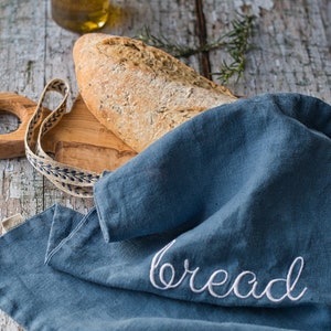May include: A rustic image featuring a loaf of bread on a wooden board, partially covered by a blue linen cloth embroidered with the word "bread". A bottle of olive oil is in the background.