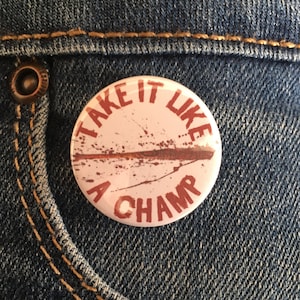 Puede incluir: Un botón redondo de color rosa claro con el texto "TAKE IT LIKE A CHAMP" en letras granates. El botón tiene un diseño desgastado con salpicaduras marrones y está colocado sobre un fondo vaquero.