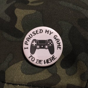 Könnte beinhalten: Ein weißer Knopf mit dem Text "I PAUSED MY GAME TO BE HERE" und einer schwarzen Controller-Grafik. Der Knopf befindet sich vor einem Tarnhintergrund.