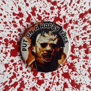 Puede incluir: Un botón redondo con el texto "PUT ON A HAPPY FACE" que rodea un retrato de un personaje de película de terror. El botón está sobre un fondo blanco salpicado de pintura roja.