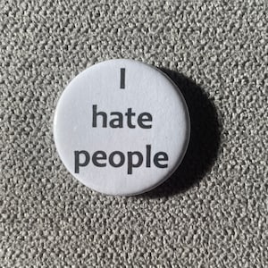 Könnte beinhalten: Ein weißer Knopf mit dem Text "I hate people" in Schwarz, zentriert auf dem Knopf. Der Knopf ist rund und liegt auf einer strukturierten, grauen Oberfläche.