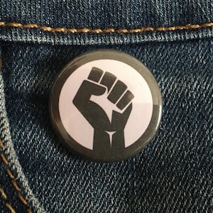 May include: A circular button badge featuring a black fist symbol on a white background, framed by a dark grey border. The badge is positioned on blue denim fabric, close to a pocket with a bronze button.