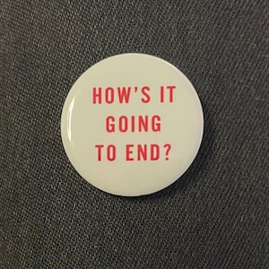 May include: A white, circular badge featuring the red text "HOW'S IT GOING TO END?". The badge has a shiny surface and is displayed against a dark grey fabric. The text is centrally aligned and clearly visible.