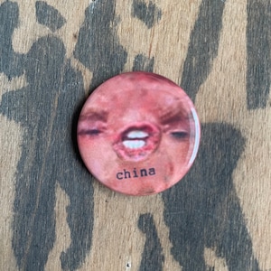 Può includere: Un bottone rotondo con un'immagine ravvicinata di un volto con gli occhi chiusi e le labbra aperte. La parola "china" è stampata in nero in basso. Il bottone è posto su uno sfondo di legno.
