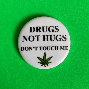 Puede incluir: Un botón blanco con el texto "DRUGS NOT HUGS DON'T TOUCH ME" en negro y un gráfico de hoja de cannabis verde. El botón está sobre un fondo verde brillante.