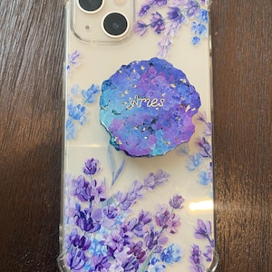 Può includere: Una custodia per telefono trasparente con un design floreale con fiori viola e blu. La custodia ha un pop socket con un design blu e viola e la parola "Aries" scritta in lettere dorate.