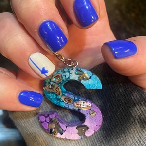 Op de afbeelding: Een handgemaakte, kleurrijke letter "S" hanger met een zilverkleurige ketting. De hanger heeft een blauw, paars en turquoise ontwerp met schelpaccenten. De nagels zijn blauw gelakt en één nagel heeft een witte basis met een blauw hart.