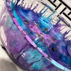 Peut inclure: Un bol en verre transparent avec un design abstrait coloré dans des tons de bleu, violet et rose. Le design présente des tourbillons et des gouttes de peinture, créant un look unique et artistique.