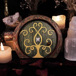 May include: A circular wooden fairy door with a green door and a gold tree design. The door features a tooth in the centre of the tree. The door is framed by brown wood with green leaves. Candles and crystals are in the background.
