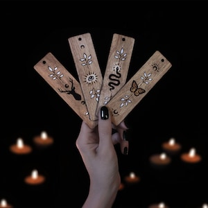 May include: Four wooden bookmarks with different designs. Each bookmark features a different symbol, including a deer, a snake, a butterfly, and a sun with an eye. The bookmarks are held in a hand against a black background.