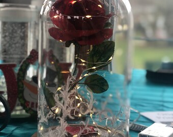 May include: A red rose with glitter is encased in a glass dome with a wooden base. The rose is surrounded by white lights and red glitter.